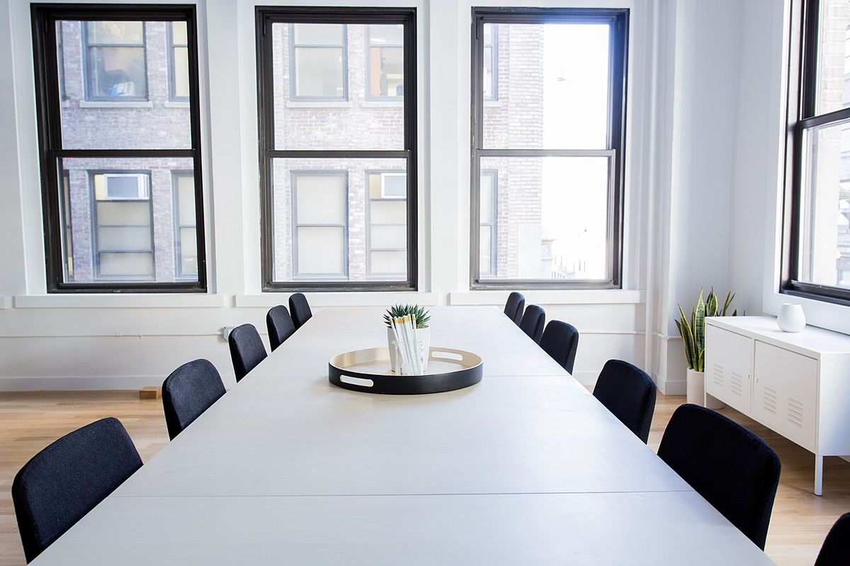 An empty conference table with chairs on both sides, in front of tall windows